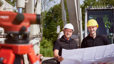 Zwei Männer mit Helm stehen auf einer Baustelle und begutachten einen Plan, den sie gemeinsam halten.