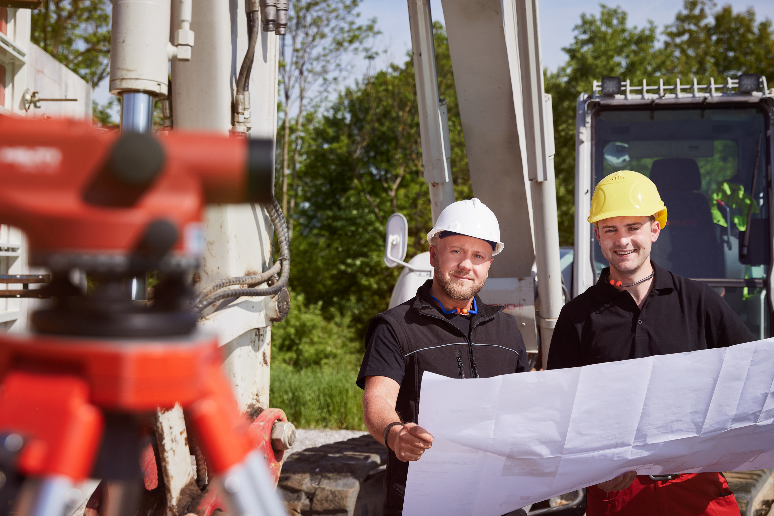 Zwei Männer mit Helm stehen auf einer Baustelle und begutachten einen Plan, den sie gemeinsam halten.