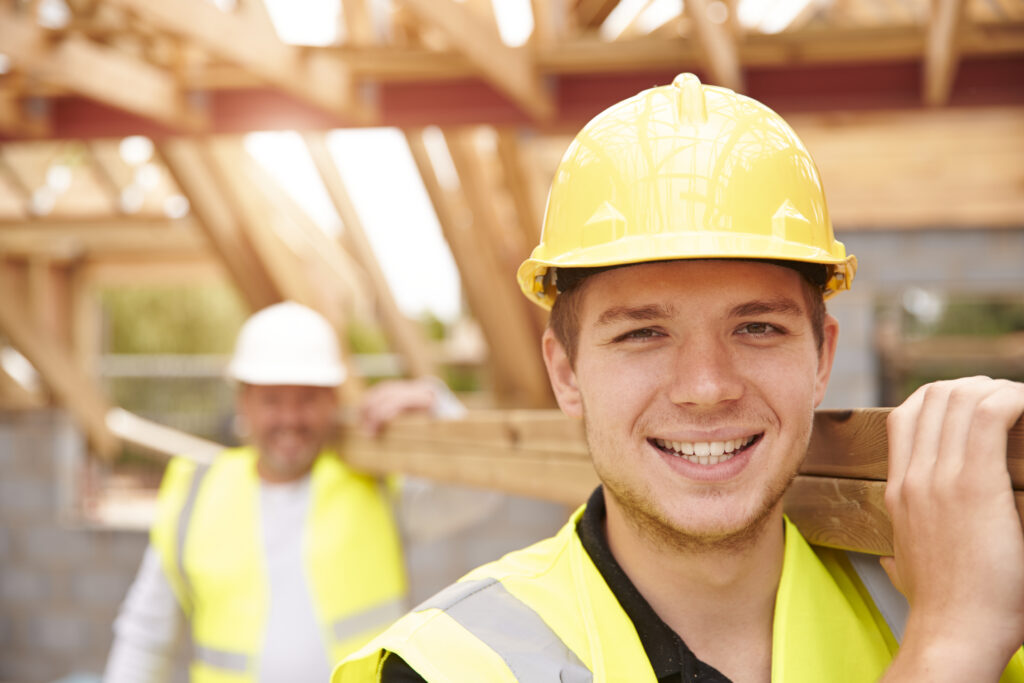 Builder And Apprentice Carrying Wood On Construction Site