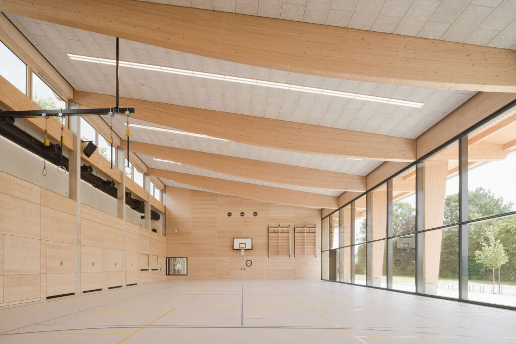Einhüftige Rahmen aus Brettschichtholz bilden im Abstand der Stahlbeton-Bestandsstützen das Tragwerk der neuen Halle. Die geschwungene Unterseite der Binder zeichnen den Momentenverlauf nach. © Brigida González