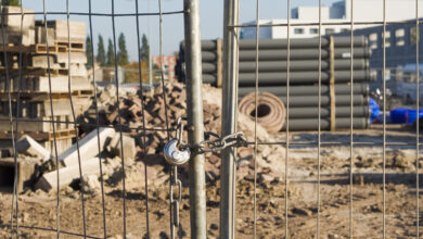 Close-up of metal fence doors which are closed with chain and padlock. In the background, out of focus, construction materals like pipes, bricks, sand, concrete. Far in the background a building and trees.