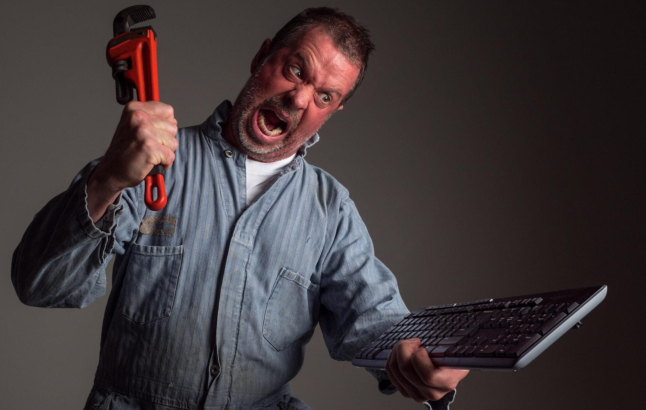 Photograph of an angry repairman hollering and about to smash a computer keyboard with an orange pipe wrench (humor).