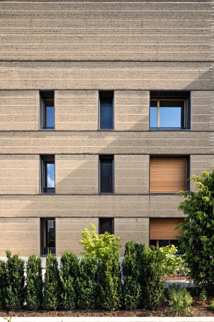 Die oberflächenfertige Stampflehmfassade wird von umlaufenden Ziegelleisten gegliedert, die zugleich als Erosionsbremsen fungieren.