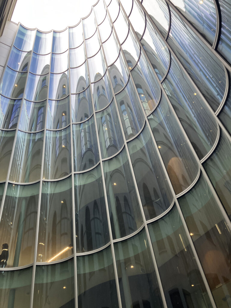 Die vertikal angeordneten, zylindrisch gebogenen Isoliergläser bilden eine durchgehende, halbmondförmige Fassadenfläche. Ihre spiegelnden Oberflächen nehmen die historische Architektur des Innenhofs auf und verstärken die räumliche Tiefe.